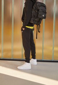 Czarna bluza z kapturem, żółty pas, czarne spodnie dresowe i białe sneakersy z wysoką cholewką. Czarny plecak z wieloma paskami. Gładka powierzchnia w tle.