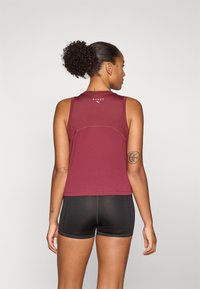 Débardeur de sport bordeaux avec une coupe ajustée, encolure arrondie et logo au dos, associé à un short noir. Texture lisse.