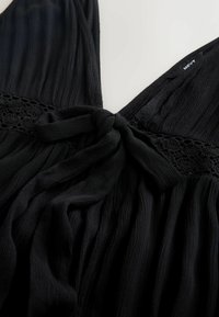 Robe en tissu noir texturé avec un nœud noué à la taille et des détails en dentelle sur la partie supérieure du milieu du corps.