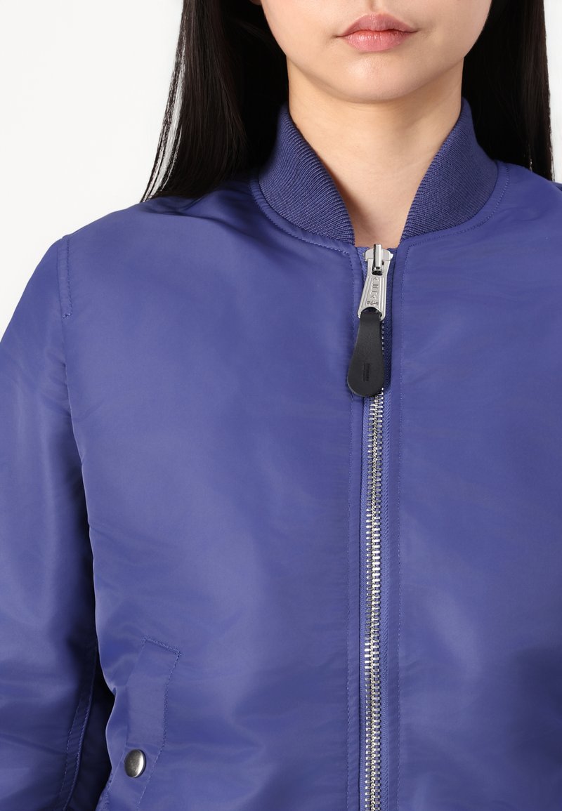 Purple bomber jacket featuring a ribbed collar, zipper closure, and a side pocket with a snap button detail. Smooth texture and casual design.