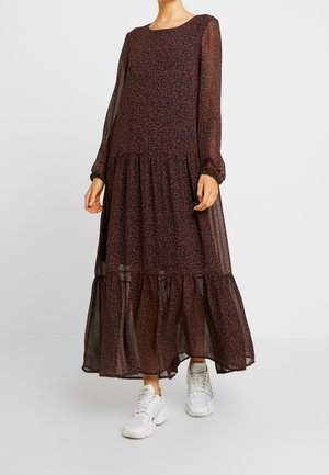 Frau trägt ein langärmliges, dunkles Maxikleid mit Blumenmuster und weiße Turnschuhe, steht vor einem schlichten weißen Hintergrund.