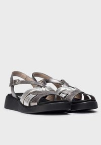 Sandalias plateadas y grises con tiras entrelazadas, acabado metálico texturizado y suela negra de plataforma. Incluye una correa ajustable en el tobillo.