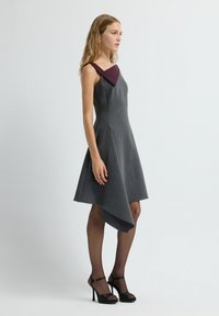 Graues Kleid mit asymmetrischem Saum, figurbetontem Oberteil und tiefmaroonem Kragen. Das Model trägt schwarze High Heels.