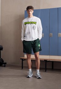 Langærmet hvid skjorte med grøn grafisk tekst, parret med grønne mønstrede shorts. Atletiske sko og en minimalistisk sølvhalskæde.