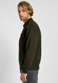 Felpa verde scuro con colletto alto, polsini a coste e chiusura con zip sul davanti. Presenta un piccolo logo sul petto. Tessuto morbido.