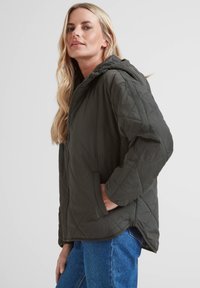 Woman with long blonde hair wearing a dark green quilted hooded jacket and blue jeans, standing in a three-quarter pose with a hand in her pocket.