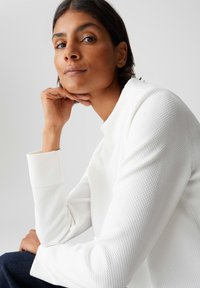 Vrouw met kort donker haar die een geometrisch wit shirt met lange mouwen draagt, leunt met haar kin op haar hand tegen een neutrale achtergrond.