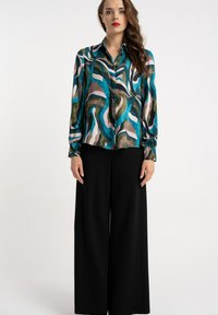 Kobieta w długim rękawie, wielokolorowej, wzorzystej bluzce oraz czarnych spodniach z szerokimi nogawkami, stojąca na jednolitym jasnym tle.