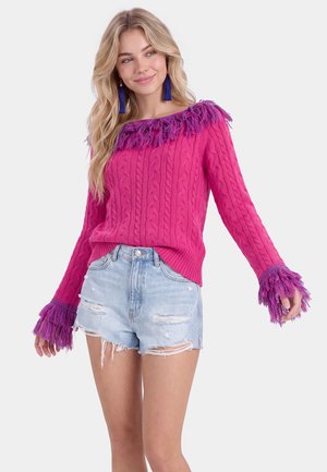 Donna che indossa un maglione rosa a trecce con frange viola sul colletto e sui polsini abbinato a shorts di denim azzurro chiaro strappati, sorridente e in posa.