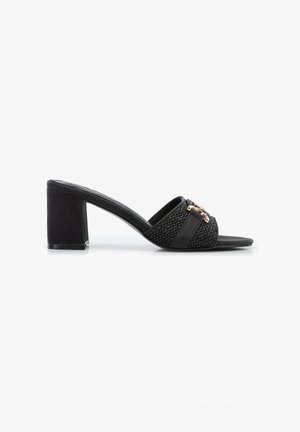 Mule nero con punta aperta, tacco largo, fascia superiore intrecciata e dettaglio a catena dorata, mostrata di profilo su sfondo bianco.