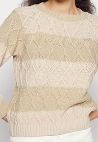 Maglione lavorato a maglia beige con un motivo a diamante, dotato di scollatura e polsini a coste e righe di diverse texture. Materiale morbido e traspirante.