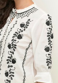 Blouse blanche avec broderie florale noire, col à volants et fermeture par lien tressé, portée par une personne aux cheveux foncés.
