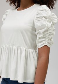 White cotton top featuring puffed sleeves with gathered detailing, a fitted bodice, and a flared hemline. Smooth texture throughout.