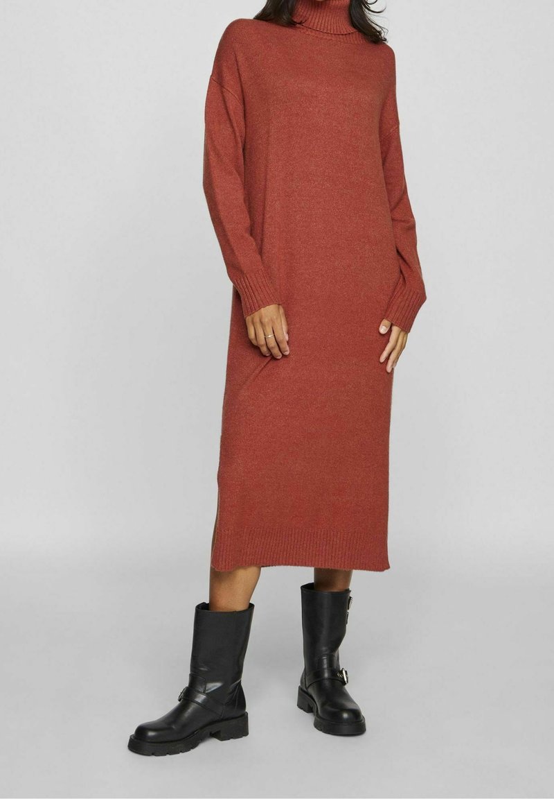 Robe en maille couleur rouille avec un col montant, des manches longues et des fentes latérales, associée à des bottines en cuir noir à talon épais.