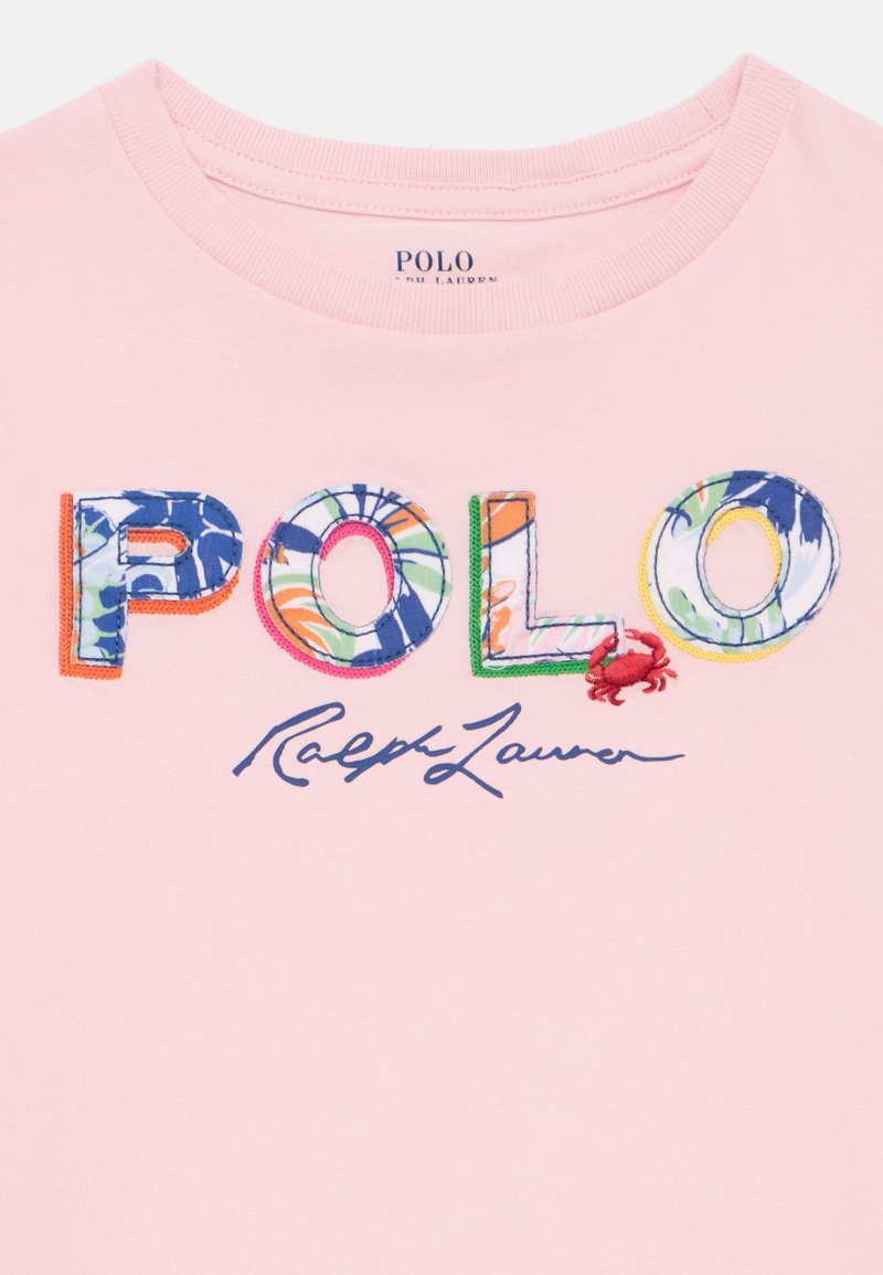 Scollatura di una maglietta rosa con lettere colorate e decorate che formano la scritta "POLO", dettaglio di un piccolo granchio rosso e firma "Ralph Lauren" sotto.