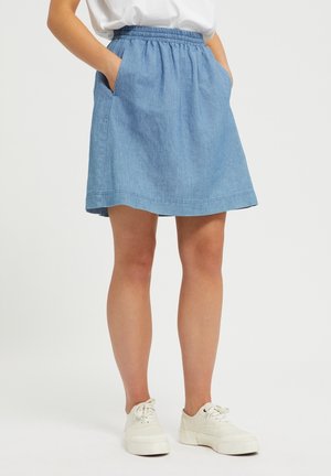 Kvinde iført en blå denimnederdel med elastik i taljen og lommer, kombineret med hvide sneakers og en hvid top, der er stoppet ned i nederdelen.