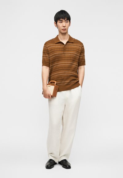 Καφέ πουκάμισο πόλο με μοτίβο και κοντά μανίκια, συνδυασμένο με φαρδιά λευκά παντελόνια. Κρατάει μια μικρή καφέ τσάντα με λαβή. Μαύρα παπούτσια.