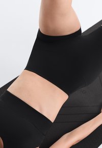 Persoon in zwarte sportkleding ligt op een zwarte yogamat, midsectie zichtbaar met armen gestrekt langs de mat.