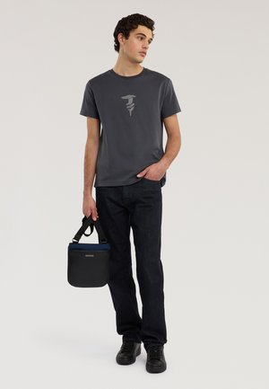 Mladý muž v tmavě šedém tričku s potiskem, černých džínách a černých teniskách, držící černou a námořnicky modrou tašku s ramenním popruhem.