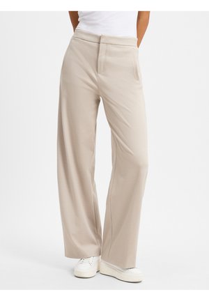 Beige Hosen mit weitem Bein und Fronttaschen, getragen mit weißen Sneakern und einem in die Hose gesteckten weißen Oberteil vor neutralem Hintergrund.