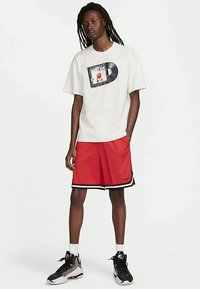 En ung person med långa dreadlocks står iförd en vit T-shirt med grafik, röda Nike-basketshorts, vita strumpor och svarta sportskor.