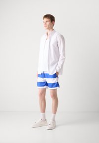 White button-up shirt over a white tank top, paired with blue and white striped swim shorts and white sneakers with socks. Minimalistic design.