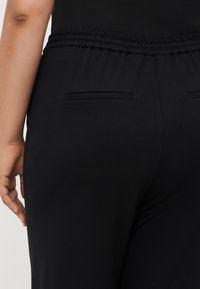 Pantalons noirs extensibles avec une taille élastique. Comprend deux poches arrière avec un design de couture subtil. Texture lisse et mate.