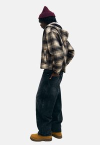 Rutig flanelljacka i bruna och krämfärgade nyanser, med huva, kombinerad med lösa svarta jeans och gulbruna kängor. Avslutad med en vinröd mössa.