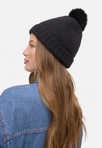 Gorro negro de punto con un acabado texturizado y un pompón de piel sintética negra en la parte superior, llevado por una persona con cabello largo y castaño.
