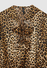 Blusa con stampa leopardata caratterizzata da colletto arricciato, texture setosa, combinazione di colori marrone e beige, e dettagli neri. Etichetta del marchio visibile.