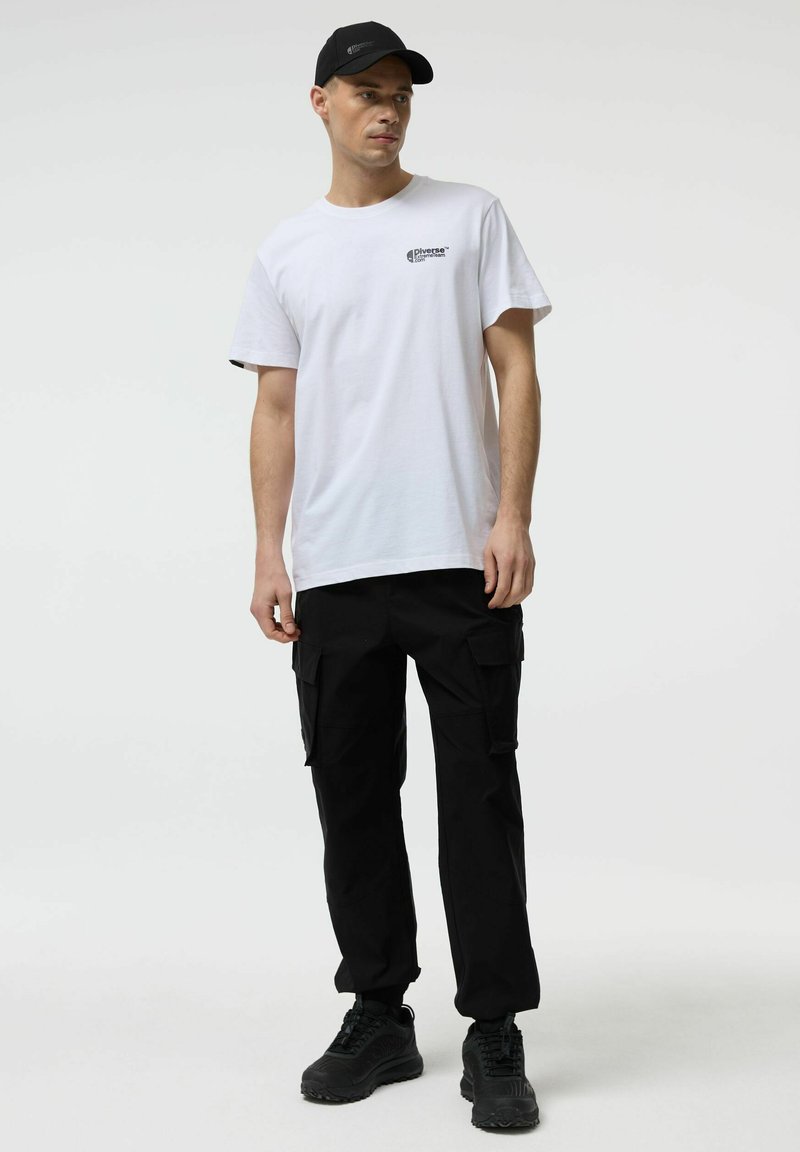 Młody mężczyzna ubrany w białą koszulkę z nadrukiem, czarne spodnie cargo, czarne trampki i czarną czapkę z daszkiem, stojący na tle jasnego, jednolitego tła.