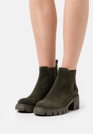 Bottines en suède vert olive avec un talon épais et une semelle crantée, dotées de panneaux latéraux élastiques pour un enfilage facile. Texture lisse dans l'ensemble.