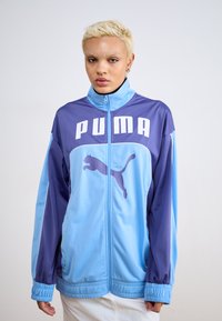 PUMA-jakke med et fargeblokkdesign i lys blå og lilla, med stor hvit logo og kattemblem, glidelås og elastiske mansjetter.