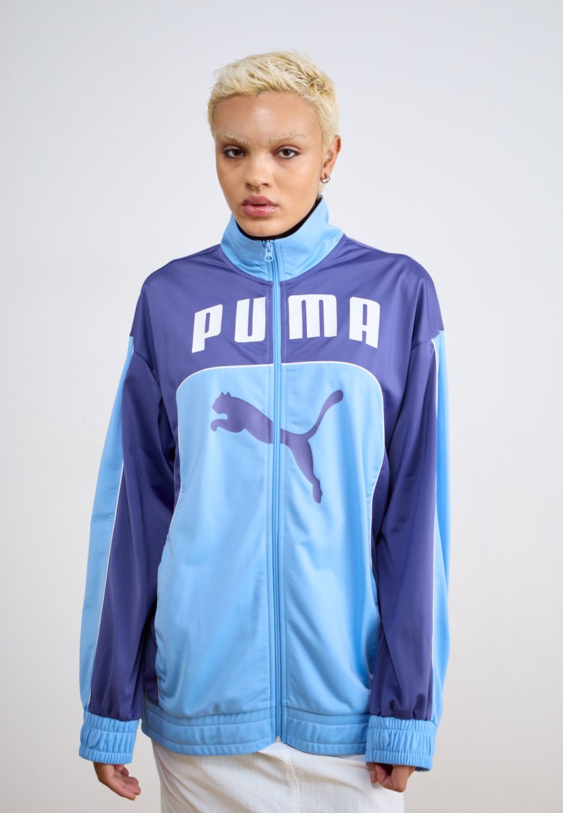 PUMA-jakke med et fargeblokkdesign i lys blå og lilla, med stor hvit logo og kattemblem, glidelås og elastiske mansjetter.