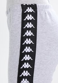 Grå träningsbyxor med en svart sidopanel som har upprepade vita logotypdesigner. Elastisk midja, mjuk textur och avslappnad passform.