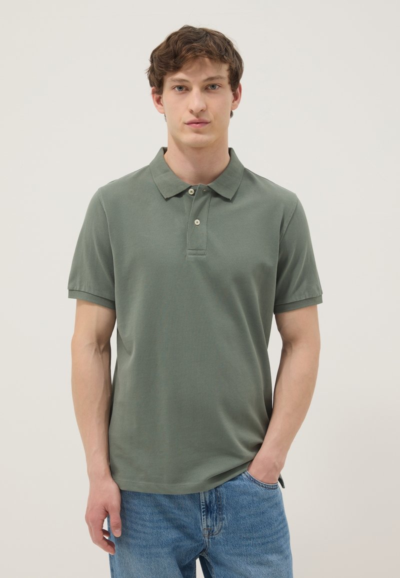 Joven con una camisa polo de manga corta de color verde oliva y vaqueros azules, de pie con una mano en el bolsillo frente a un fondo sencillo.