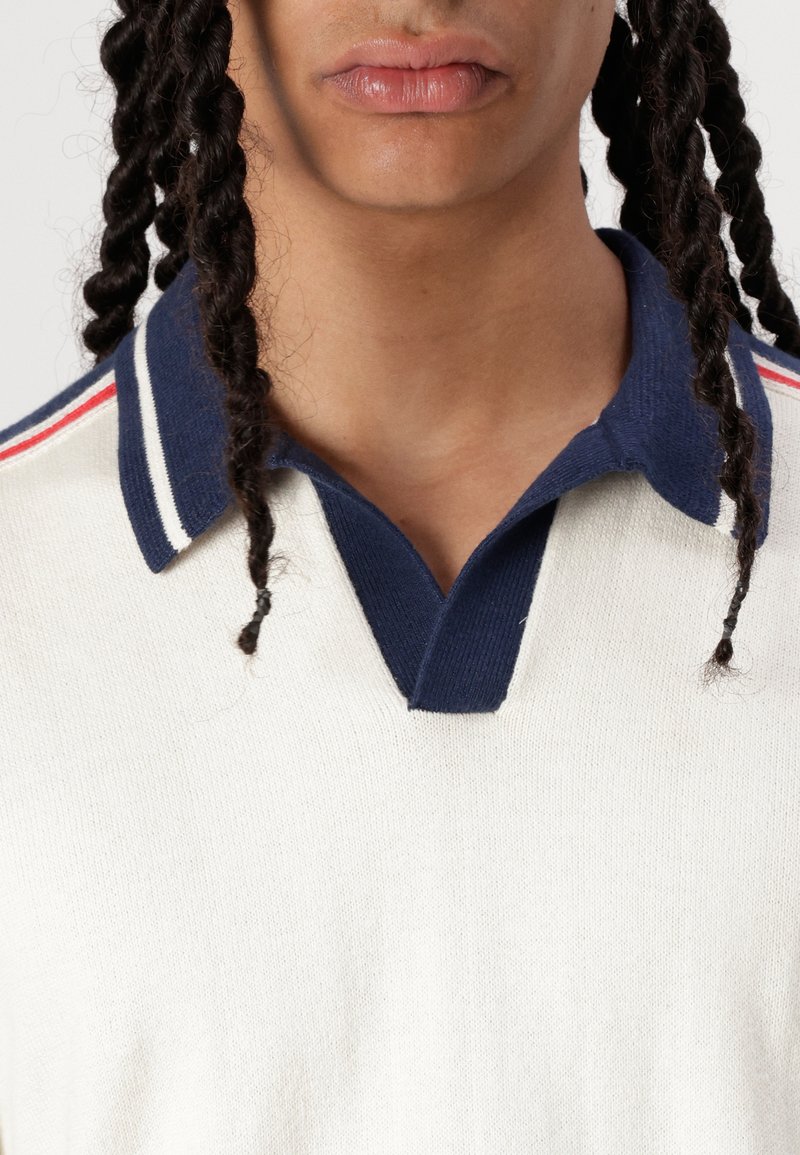 Camisa polo color crema con cuello azul marino y rayas rojas y blancas. Tejido texturizado, con diseño de cuello en V.