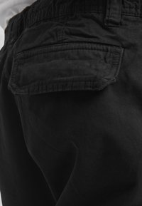 Gros plan sur un pantalon noir avec un rabat de poche arrière et une taille élastique, montrant la texture du tissu et les détails de couture.