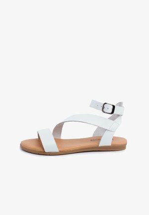 Sandalia de cuero blanca con diseño de tiras cruzadas, hebilla en el tobillo y una suela plana en tono marrón con acabado texturizado.