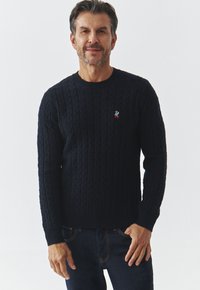 Marineblauwe gebreide trui met een gestructureerd kabelontwerp, ronde hals en een kleine kleurrijke logo-accent op de borst. In combinatie met donkere spijkerbroeken.