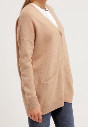 Femme portant un cardigan en tricot beige ample par-dessus un top blanc et un jean bleu, debout devant un fond clair uni.