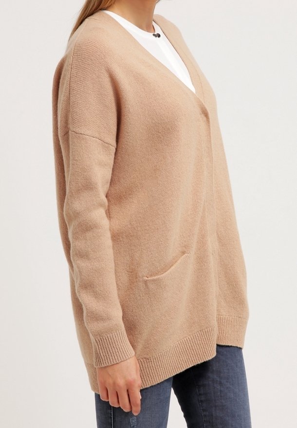 Femme portant un cardigan en tricot beige ample par-dessus un top blanc et un jean bleu, debout devant un fond clair uni.