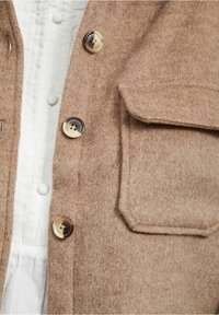 Veste en feutre marron avec de gros boutons, une poche plaquée et une texture douce. Portée par-dessus une chemise blanche à boutons.