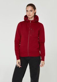 Sudadera roja con cremallera, capucha ajustable y puños acanalados, además de un bolsillo con cremallera en el pecho. Textura suave y diseño ajustado. Detalles en negro.