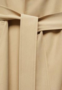 Beżowy materiał z detalem w postaci przewiązanego pasa, teksturowana powierzchnia oraz precyzyjne szwy. Projekt obejmuje dopasowane fałdy i czyste linie.