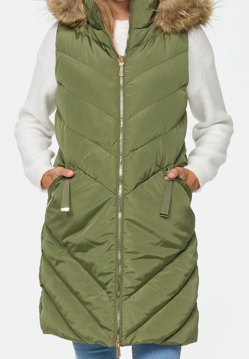 Personne portant un gilet matelassé sans manches vert à capuche avec bordure en fourrure par-dessus un pull blanc à manches longues, mains dans les poches du gilet.
