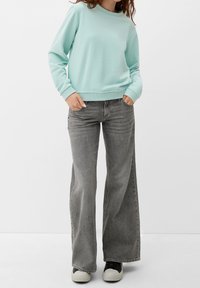 Mujer con un sudadera azul claro, vaqueros de pierna ancha gris descoloridos y zapatillas negras y blancas, de pie con las manos en los bolsillos.