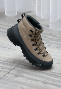 Beige og sorte vandrestøvler med tekstureret sål og robuste snørebånd, placeret på en glat marmoroverflade med lys, der filtrerer igennem.
