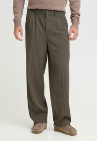 Donker groene, geplooide broek van gestructureerde stof, met een knoopsluiting aan de voorkant en zijzakken, gecombineerd met bruin, zacht-soled schoenen.