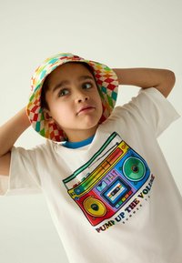 Biała koszulka z kolorową grafiką boomboxa, niebieskim akcentem przy kołnierzyku, w połączeniu z kratkowaną czapką typu bucket w jaskrawym czerwonym, zielonym i żółtym kolorze.